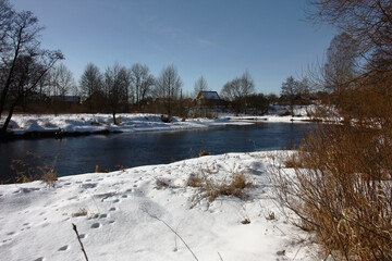Sunny winter day. Minsk region of Belarus. Between snow-covered coast river Bobr water flows. Rural houses are visible in the distance behind turn of the river. © imamchits