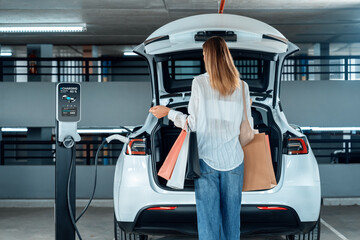 Young woman travel with EV electric car to shopping center parking lot charging in downtown city...