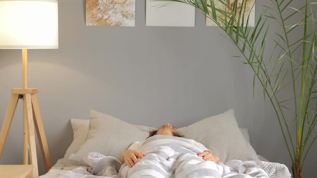 Sleepy morning. Brown-haired girl wearing white T-shirt sitting in bed after early waking up with lack of energy getting down again needs more time for rest.