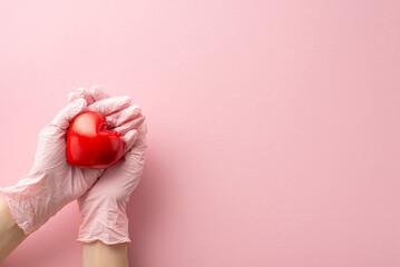 Health concept. First person top view of my hands in surgical gloves, gently grasping a red heart, a symbol of an organ, on a pastel pink surface with room for text or promotions