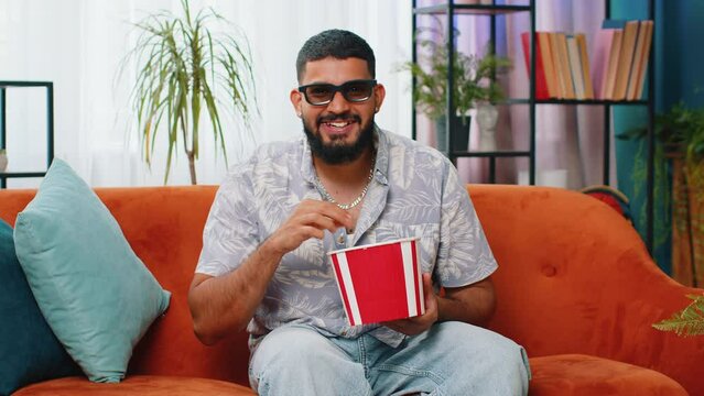 Excited Indian man sitting on sofa eating popcorn and watching interesting tv serial, sport game, film, online social media movie content at home apartment. Guy enjoying domestic entertainment indoors