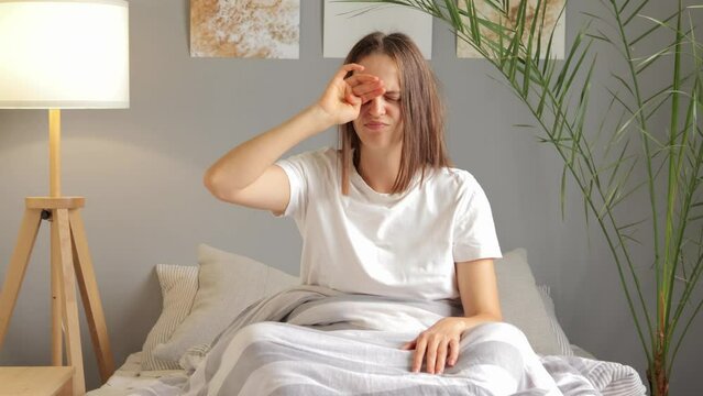 Tired sleepy woman wearing white T-shirt rubbing eyes with her hand while sitting on bed waking up in bad mood being late for work having headache because of lack of sleep.