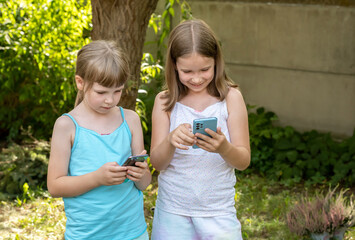 Girls children holding their smartphones in hands, kids outdoors looking at their mobile phones laughing, using technology, apps, outside scene, real people lifestyle. Tech savvy young kids concept