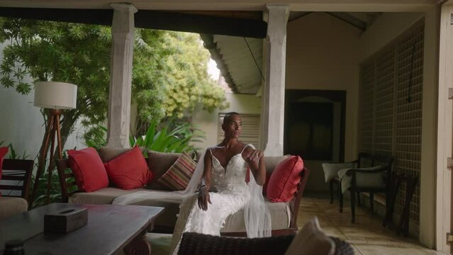 Non-binary black bride in wedding dress sits, poses in living room on sofa of retro vintage villa. Elegant non-binary ethnic person smiles at camera in stylish interior of luxury hotel.