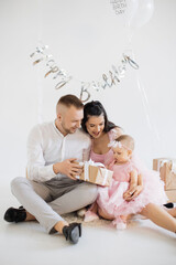 Little baby girl with her positive young parents sitting in studio decorated with balloons and unwrapping birthday present. Loving caucasian family of three having fun during anniversary celebration.