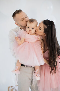 Cheerful young parents kissing little daughter wearing festive pink dress and creating precious memories during first anniversary in white studio. Concept of love, holiday and celebration.
