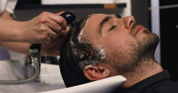 A handsome young man with a smile is resting in a hairdressing salon. After dyeing the hair brown, the hairdresser thoroughly washes the young man's head with water. Slow motion.