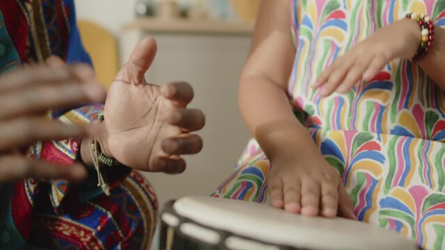 Close up shot of cheerful African American father in dashiki kaftan and kufi hat clapping hands as his daughter playing djembe drum on Kwanzaa home celebration