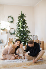 Obraz na płótnie Canvas Cheerful young parents sitting on floor near shiny Christmas tree and embracing cute daughter on toy sledge. Caucasian family of three enjoying festive activities at cozy home.