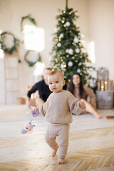 Focus on adorable baby girl in knitted wear running with barefoot at cozy apartment with festive decor. Joyful young parents sitting near Christmas tree on blurred background. Winter holidays concept.