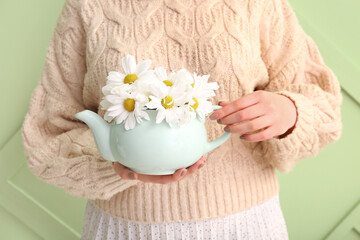 Beautiful young woman with teapot of chamomile tea and flowers near green wall