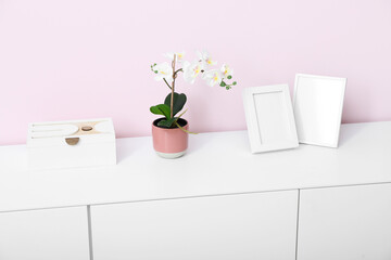 White chest of drawers with orchid flower and blank pictures near pink wall in room, closeup