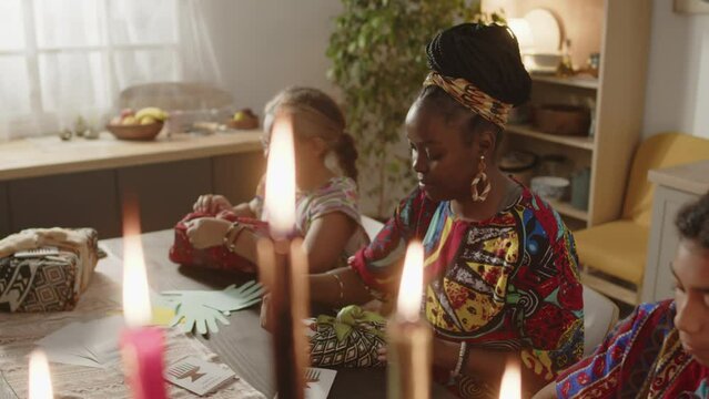 High angle shot of African American woman in dashiki kaftan and headwrap talking to little daughters while wrapping gifts for family on Kwanzaa holiday