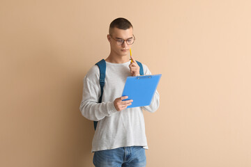 Thoughtful male student in eyeglasses with clipboard on beige background