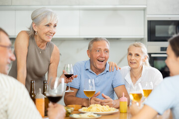Happy carefree older man and women gathering around kitchen table, sipping alcoholic drinks with snacks, cheerfully chatting, laughing and sharing stories. Seniors communication concept