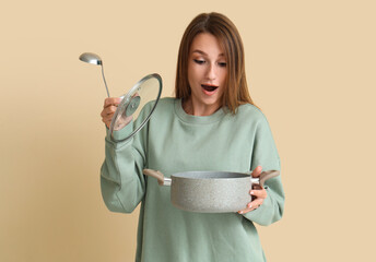 Surprised young woman with ladle and cooking pot of chicken soup on beige background