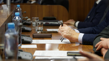 A man in a suit holds glasses in his hands next to a document while sitting at a table next to his colleagues. Official, lawyer, or businessman. Photo. Selective focus. No face