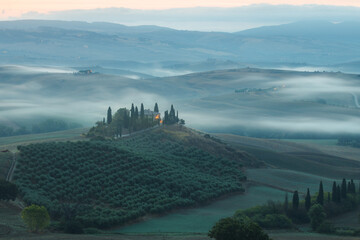 Moody, atmospheric early morning mist in the valley of Val d'Orcia and a rustic farmhouse in the scenic countryside landscape and rolling hills of rural Tuscany, Italy.