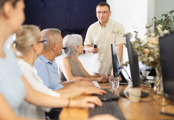 Enthusiastic male tutor teaching group of seniors in computer workshop, promoting technology...