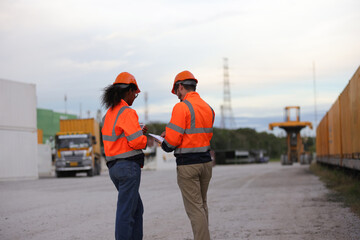 Male and Female logistic  engineers with orange safety jackets hold tablets working at the sideline of the Container Yard