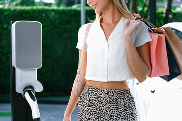 Young woman travel with EV electric car charging in green sustainable city outdoor garden in...