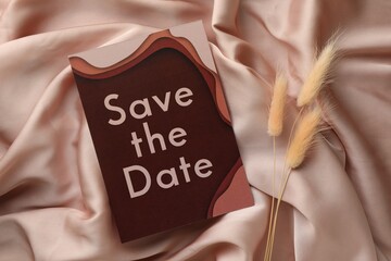 Beautiful wedding invitation card with Save the Date phrase and dry flowers on pink fabric, flat lay