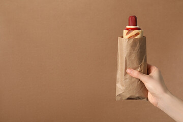 Woman holding paper bag with delicious french hot dog on brown background, closeup. Space for text
