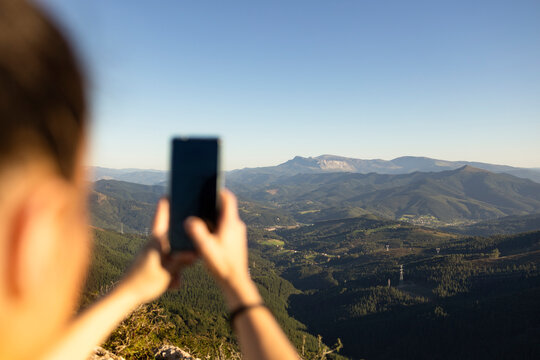 A happy young woman takes pictures of the landscape with her smartphone from the top of a mountain.