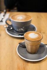 A grey coffee cup and saucer of flat white coffee, and a smaller piccolo in a glass (both with heart latte art) on a wooden table at a cafe in Cairns — Far North Queensland, Australia