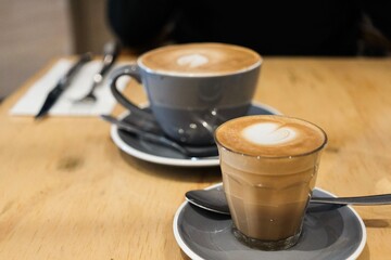A grey coffee cup and saucer of flat white coffee, and a smaller piccolo in a glass (both with heart latte art) on a wooden table at a cafe in Cairns — Far North Queensland, Australia