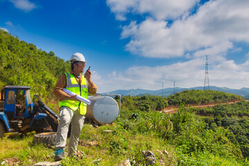 Engineer manager man with white helmet and hold construction paper plan and fm handheld transceiver or walkie talkie look back to camera and stand in front of mountain.