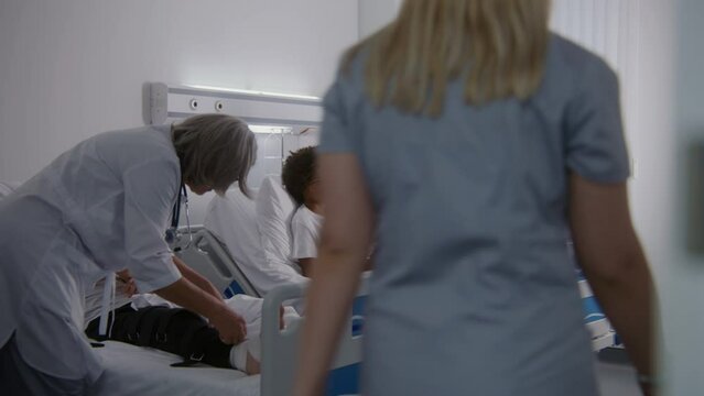 Mature physician bandages leg of young boy. African American man visits son in hospital ward. Nurse comes to teenager and raises back of his bed. Patient fully recovering after successful operation.