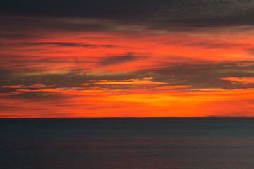 Clouds over the ocean being lit by the sun from below the horizon. The clouds are orange, red and yellow, and the sea is still very dark.