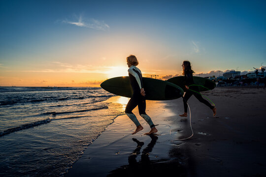 Two surfers with surfboard running at beach preparing to hit the waves at sunset.