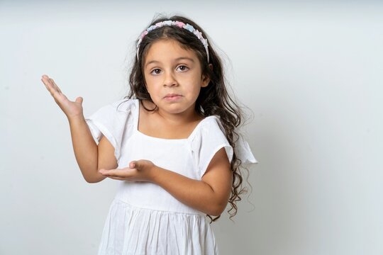 beautiful kid girl wearing white dress pointing aside with both hands showing something strange and saying: I don't know what is this. Advertisement concept.