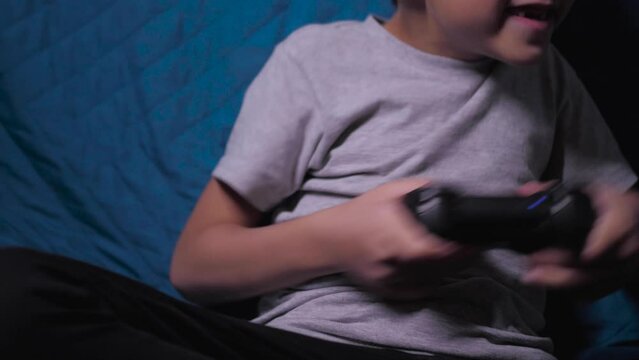 child a boy of six years old is sitting on the couch at home in the evening and playing video games with a black joystick