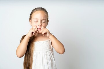 Pleased beautiful kid girl wearing white dress with closed eyes keeps hands near cheeks and smiles tenderly imagines something very pleasant