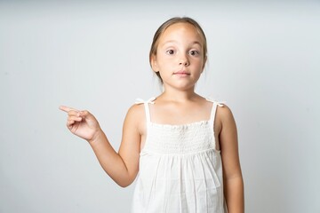 Positive beautiful kid girl wearing white dress with satisfied expression indicates at upper right...