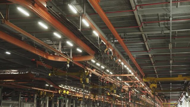 Guide rails under ceiling of assembly shop with lifting mechanisms and many tubes for pneumatic impact wrenches. Large production with lights and hoists on in factory under roof.