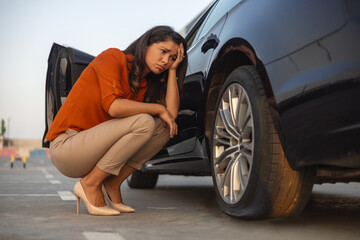 Portrait of a despair woman crouching next to her car with flat tire, problems on the road,...