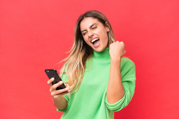 Young Uruguayan woman isolated on red background with phone in victory position