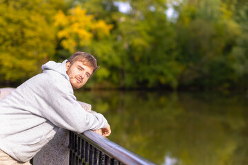 attractive young man, handsome guy in autumn weather in park, on embankment to river, sunbathing outside in foliage in sunset, breathing fresh air in sun. male portrait in fall. copy space, text