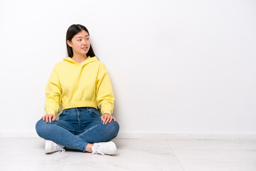 Young Chinese woman sitting on the floor isolated on white wall looking side