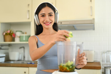 Obraz na płótnie Canvas Young woman in fitness clothes making green detox vegetable smoothie with blender in kitchen. Dieting, healthy eating concept