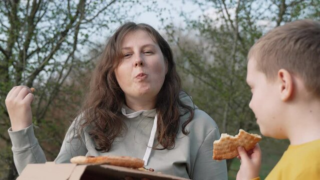 Caucasian plus size woman demonstratively, defiantly, teasingly eats slice of pizza at camera in park. Concept of quitting diets, accepting your body. Mother, son had picnic with fast food in nature.
