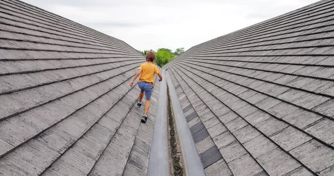 Young boy fearlessly runs, jumps, and explores rooftop, displaying agility and playfulness. Slow motion footage captures his daring movements as he navigates roof slopes and jumps over gutter.