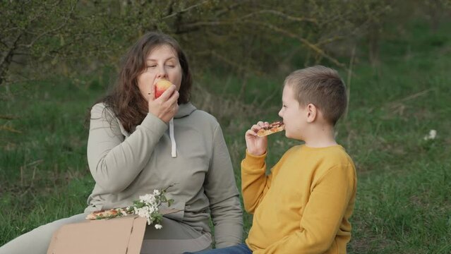 Mother and son chew food of contrasting healthiness during picnic in park. Plus size woman bites fortified fresh apple while following diet. Boy chews greasy pizza on free diet.