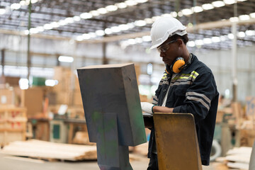 African American male engineer working and using laptop computer maintenance or inspecting quality of machine for manufacturing lines in wooden warehouse storage plant