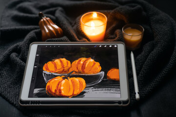 The still life picture with pumpkins drawn on the electronic tablet next to burning candle and ceramic pumpkin figurine on black table. The concept of inspiration, creativity, self-development, hobby,