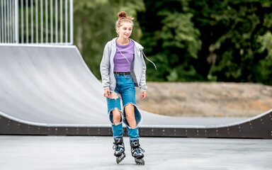 Beautiful girl roller skater riding in city park with ramp. Pretty female teenager rollerskating in...